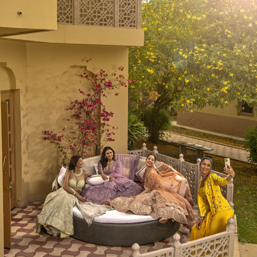 A group of women enjoying tea in a cozy garden setting with lush greenery, comfortable pillows, and ornate furniture at dusk.