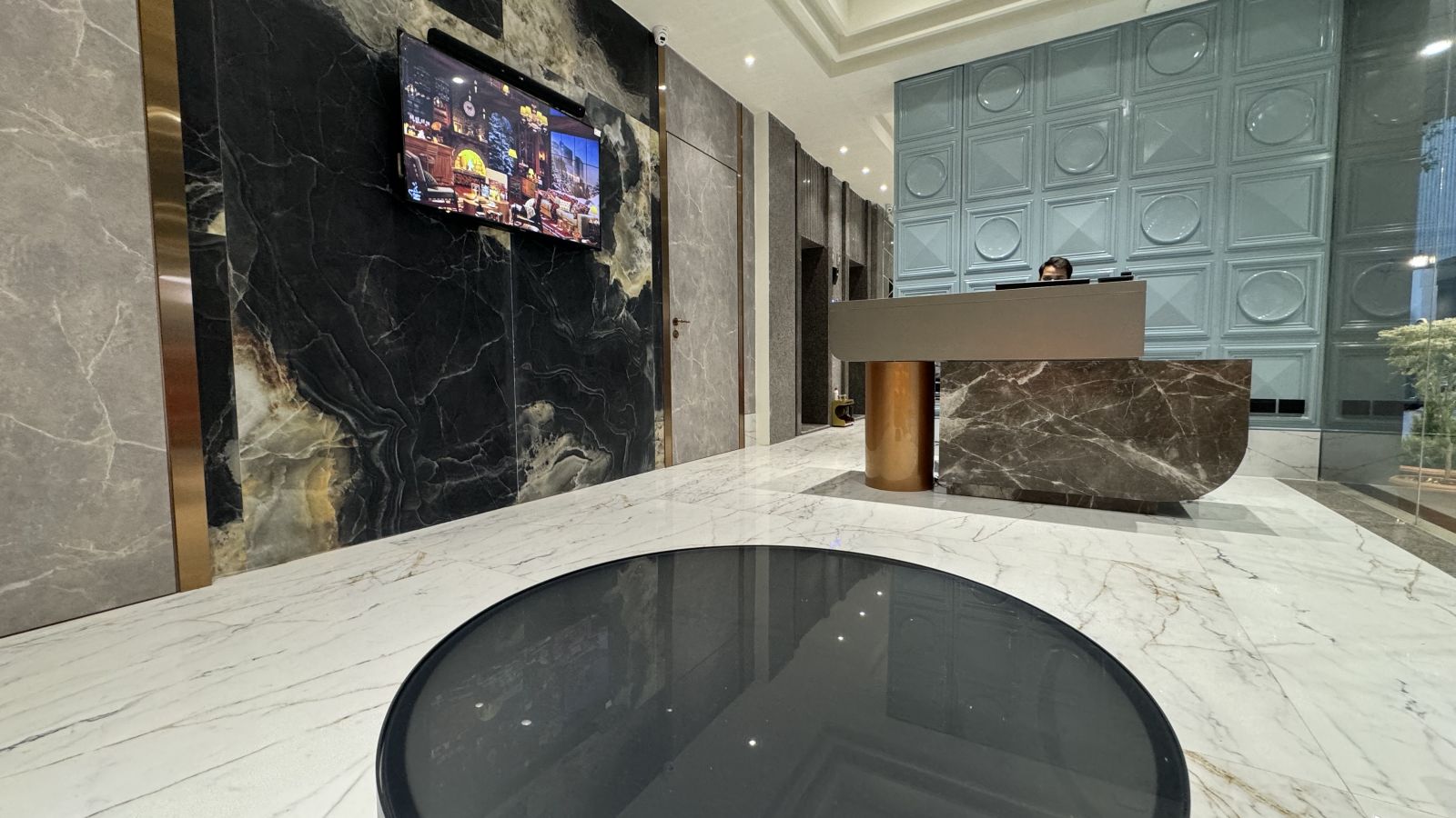 The reception area of Hotel Mumbai House, featuring a large marble desk with a wooden front, a large-screen TV mounted on a black marble wall, and a patterned ceiling with downlights.