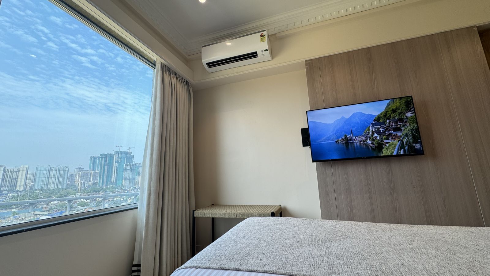 A bright hotel room with a large window offering a city view, a wall-mounted TV, a bed with a gray headboard, and a wooden side table.