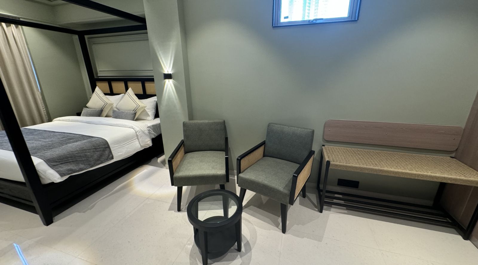 A modern hotel room featuring a black four-poster bed with white bedding and decorative pillows, two gray armchairs, a black round coffee table, and a wooden bench against a wall with a high window.