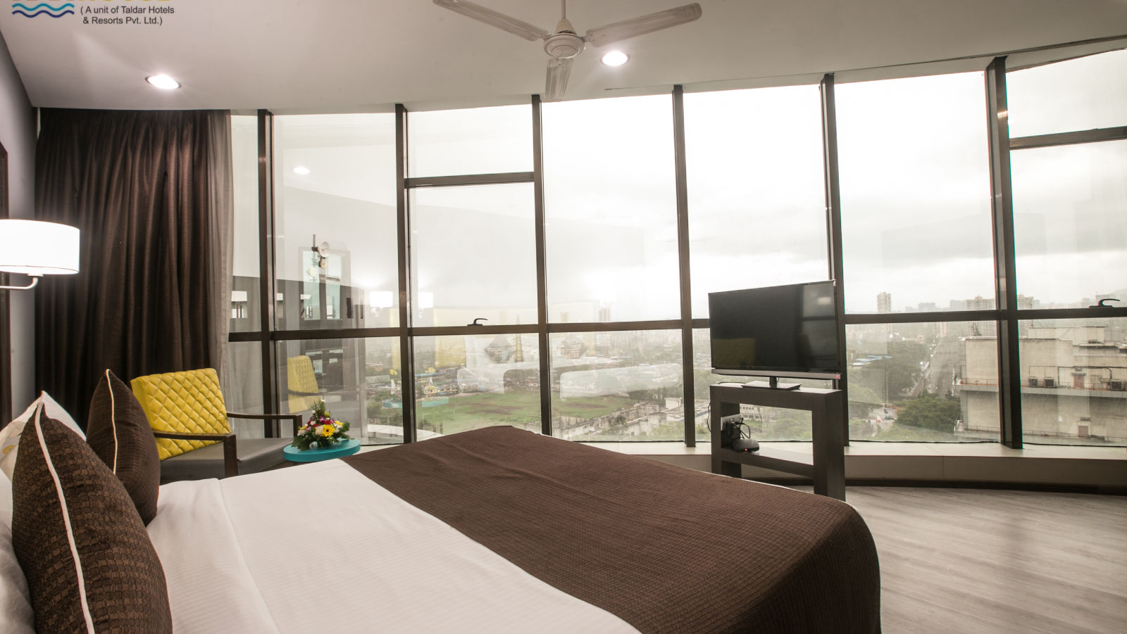 Hotel room with a large window offering a city view, a bed with brown bedding and decorative pillows, and a flat-screen TV.