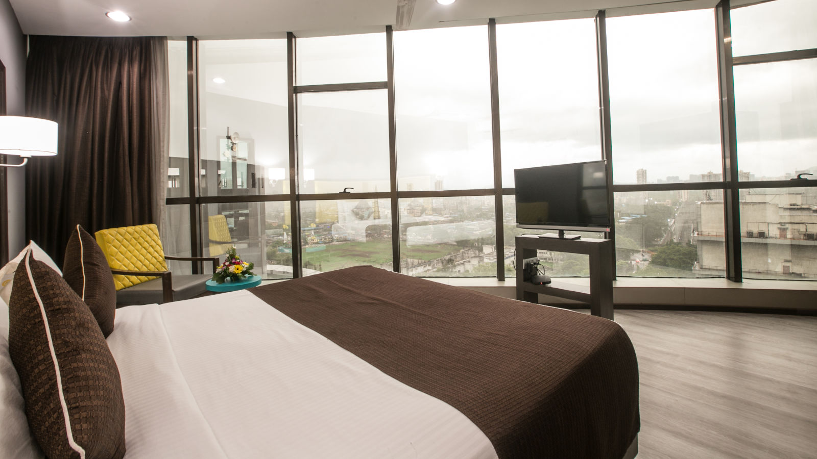 Hotel room with a large window offering a city view, a bed with brown bedding and decorative pillows, and a flat-screen TV.