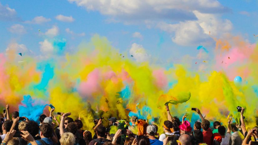 a crowd of people playing holi and throwing colours in the air with blue sky in the background