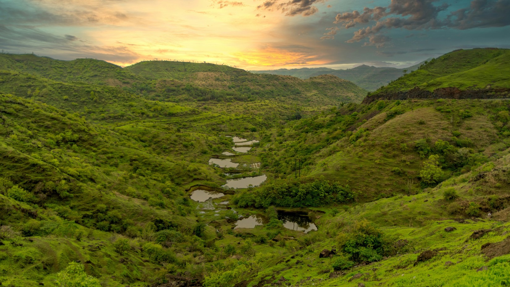 A panoramic view of a lush green valley dotted with water bodies at sunset, showcasing a dramatic sky painted with warm hues.