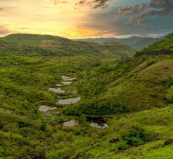 alt-text A panoramic view of a lush green valley dotted with water bodies at sunset, showcasing a dramatic sky painted with warm hues.