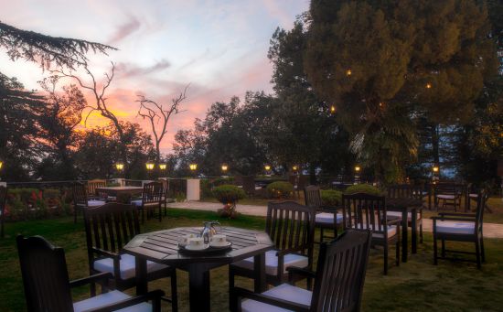 alt-text An outdoor dining space with multiple wooden chairs and tables, surrounded by lush green trees.