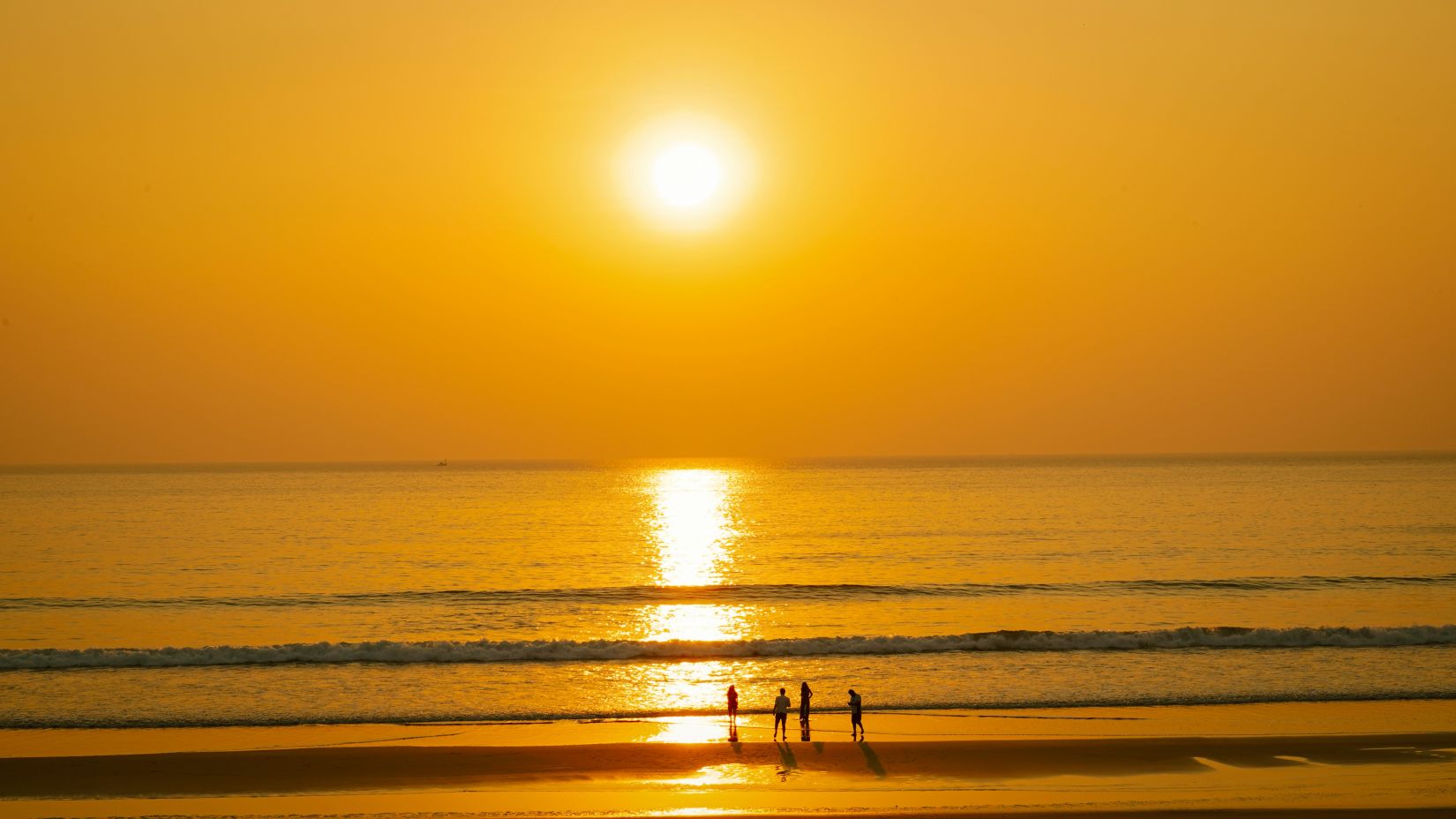 A far out view of the beach with people standing on the shores and the sun setting in the background