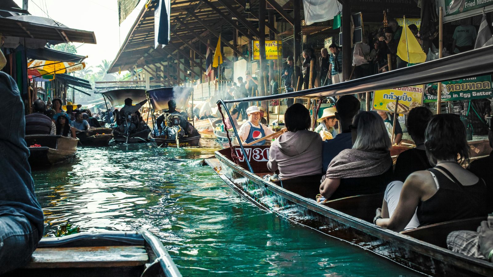 many people sitting in a boat travelling through a bustling market