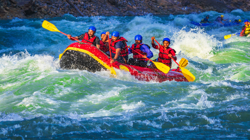 A raft filled with seven people, wearing red life jackets and helmets, is navigating through turbulent, white-capped waters. Everyone appears exhilarated by the action.