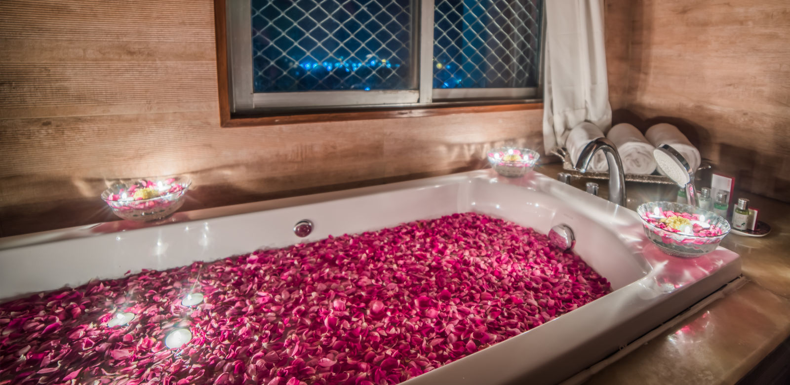 A romantic setting with a bathtub filled with rose petals and surrounded by candles, with a window overlooking the night sky -Sitara - Luxury Hotel