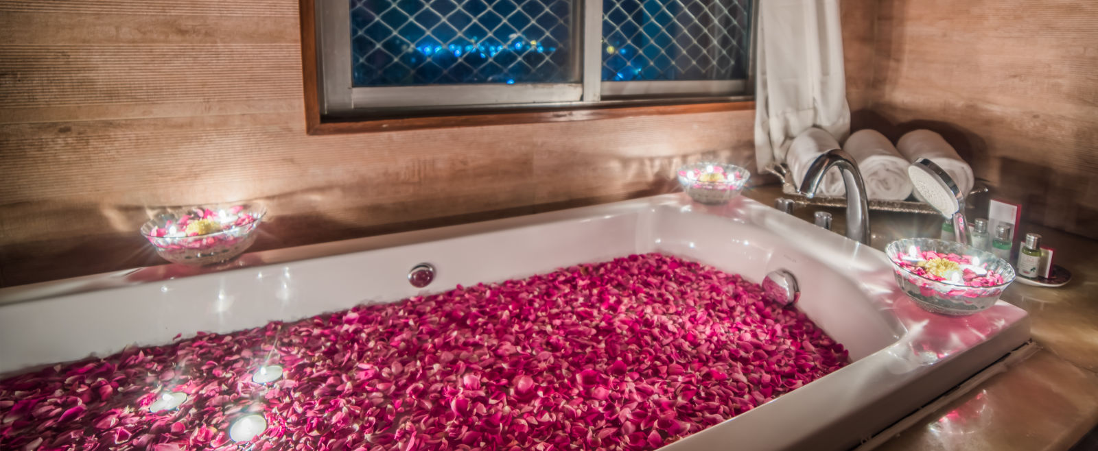 A romantic setting with a bathtub filled with rose petals and surrounded by candles, with a window overlooking the night sky -Sitara - Luxury Hotel