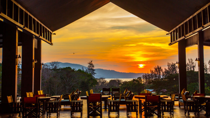 Wooden chairs in an open restaurant that overlooks a setting sun - Symphony Samudra Beachside Jungle Resort And Spa