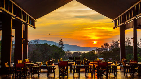 Wooden chairs in an open restaurant that overlooks a setting sun - Symphony Samudra Beachside Jungle Resort And Spa