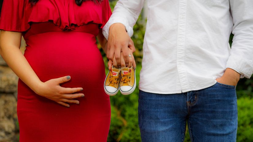 A pregnant woman hold her belly as she stands next to her partner the latter holds up a pair of baby shoes -Babymoon Near Delhi