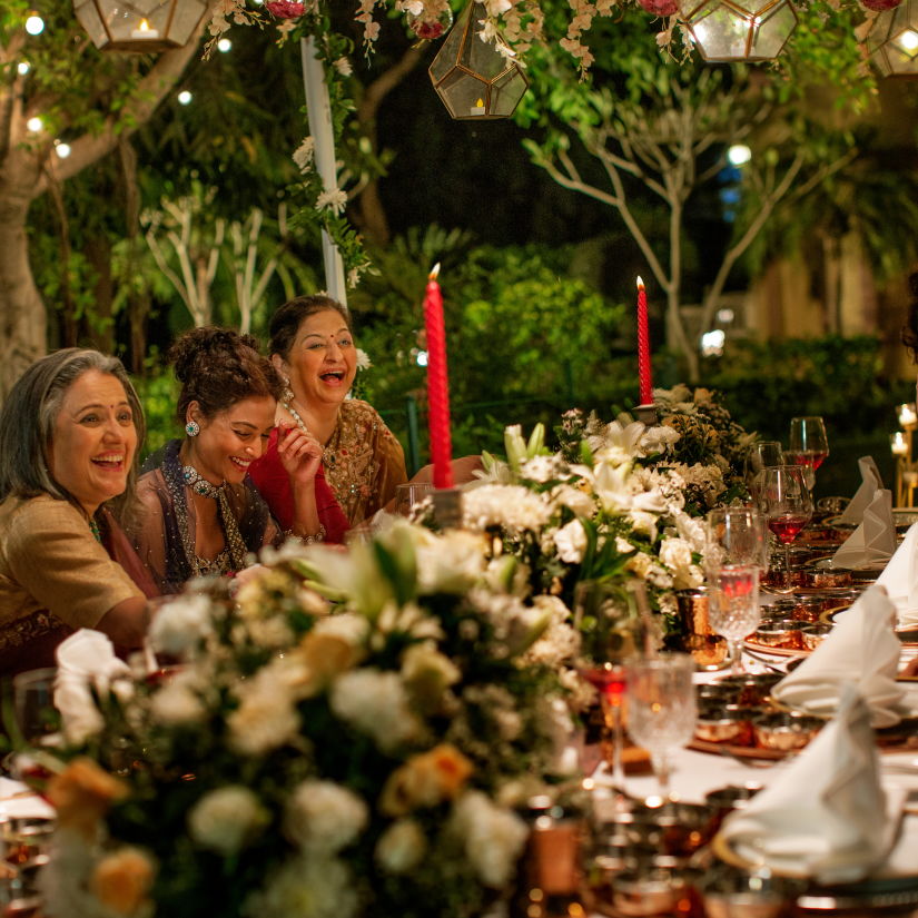 An outdoor evening dinner party with people laughing around a table decorated with candles and floral arrangements, strung lights above adding a warm glow.