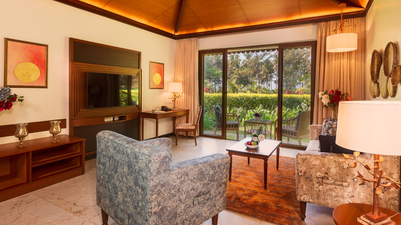 Gamyam Retreat, Kumta - The living area with sofas and television and a ceiling to door window opening to a garden