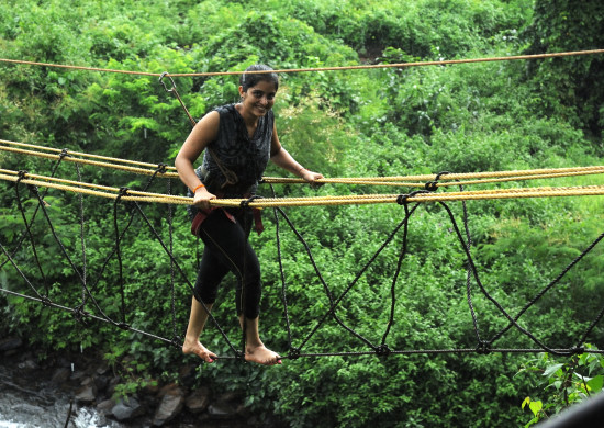 alt-text A focused individual crossing a rope bridge over a lush, green area. They appear determined, showcasing an adventurous outdoor activity.