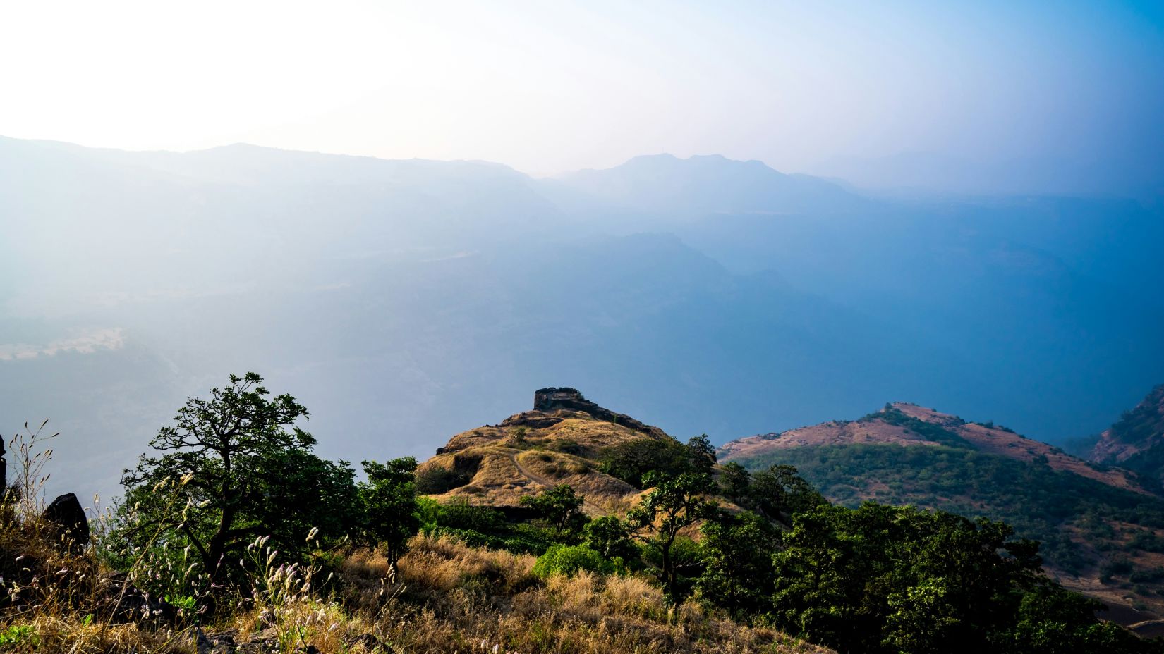  A serene mountain vista bathed in the soft glow of a hazy sunrise with clear blue skies above. - Fort Jadhav GADH