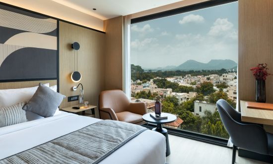 alt-text Side view of a spacious room ,  featuring a large bed, full-length windows, and a chair-table set at Parallel Hotel Udaipur