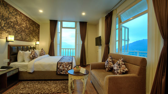 full view of a lavish bedroom with wide open windows and  a balcony along with a couch in front of the window and teapoy with a fruit basket placed in front of the couch - the aryan regency