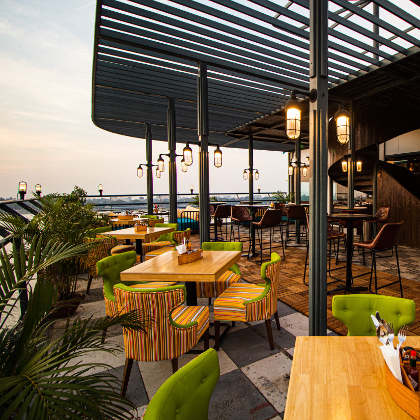 A scenic rooftop restaurant with colorful chairs, wooden tables, and a view of the sunset over a river.