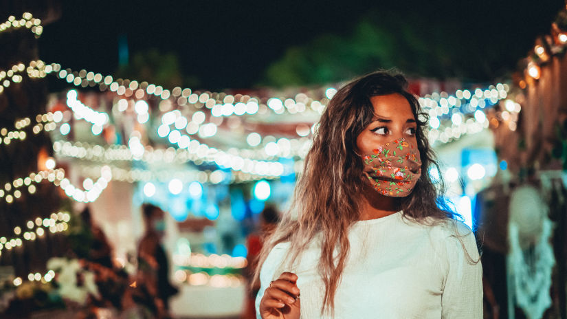 A low depth mid shot of a woman wearing mask with bright lights of a market in the background taken at night