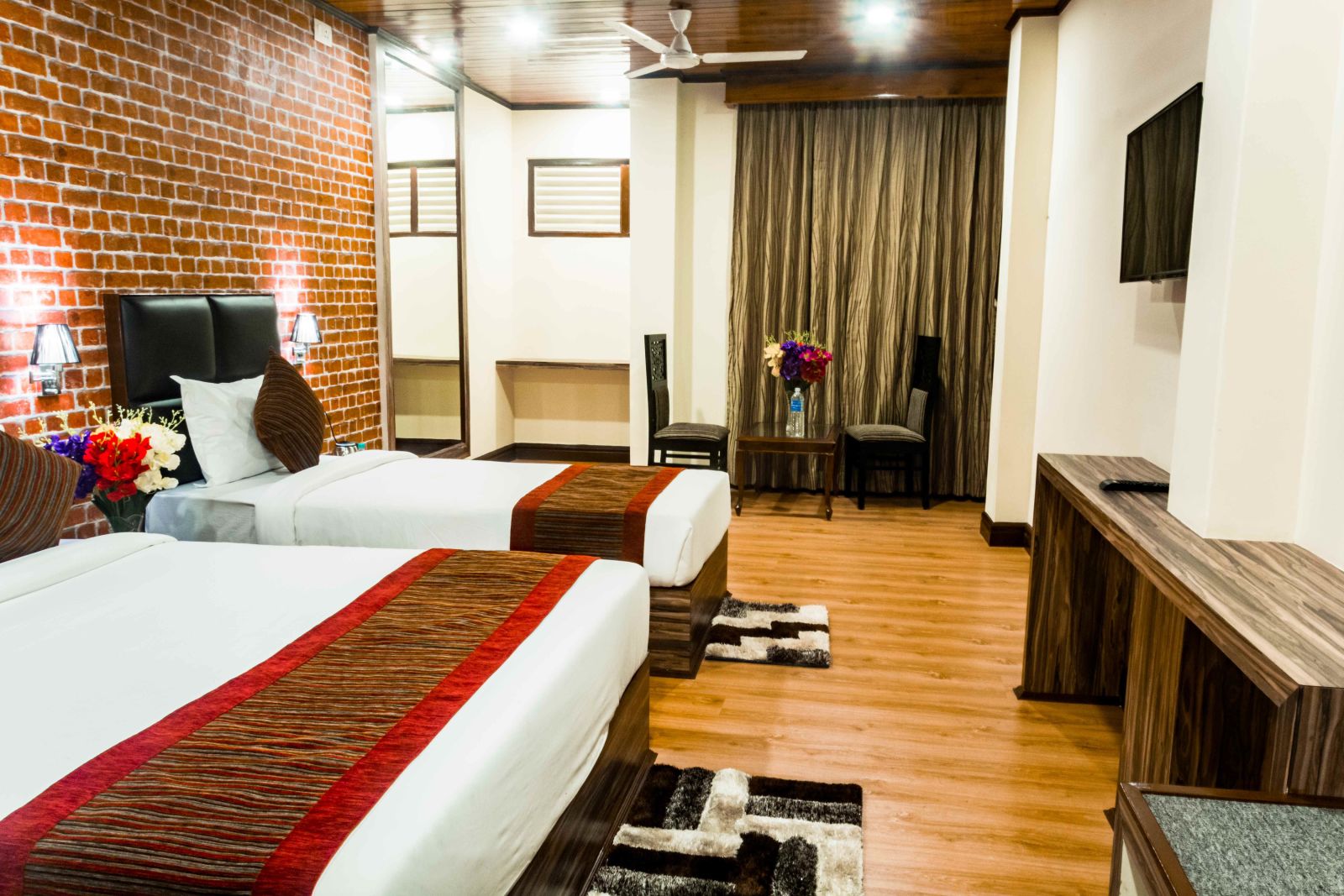 An image of the premium room with twin beds, bed side tables, flat screen TV, chairs and table, and floral decoration with bright lighting - Ecotel Tin Tin, Bijanbari