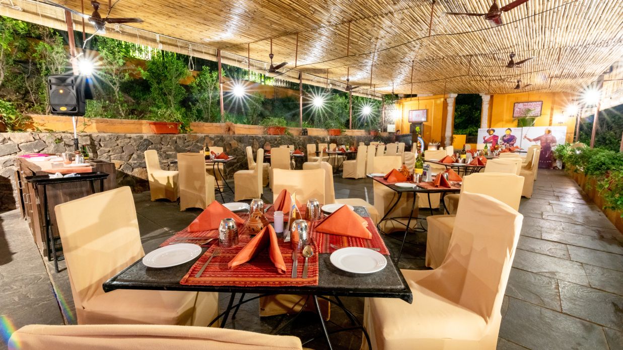 An open-air restaurant with thatched roof, cream upholstered chairs, and tables set with red napkins  at Fort JadhavGADH 