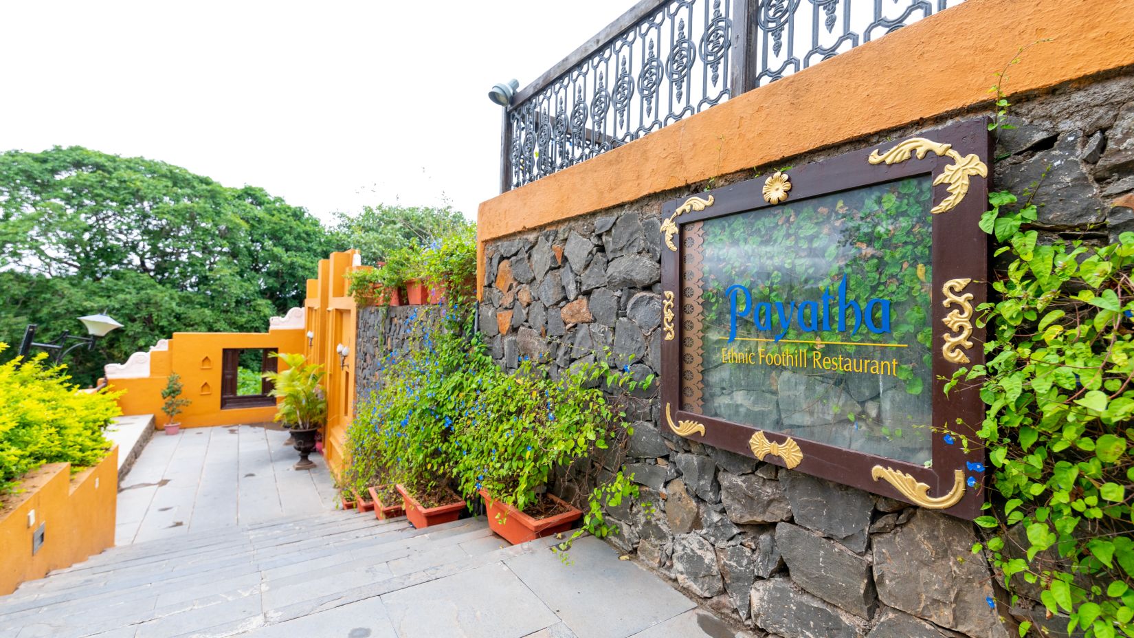  A pathway leading to a vibrant orange entrance with lush greenery and a stone wall bearing a hotel sign -Fort JadhavGADH 