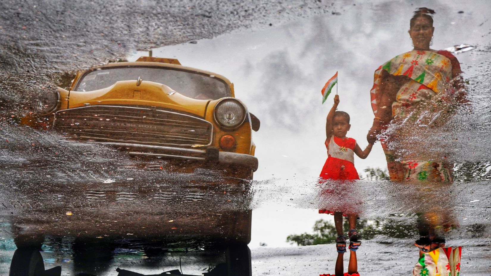 an image of a yellow Kolkata taxi and a little girl with her mother walking next to it while the girl is holding the indian flag