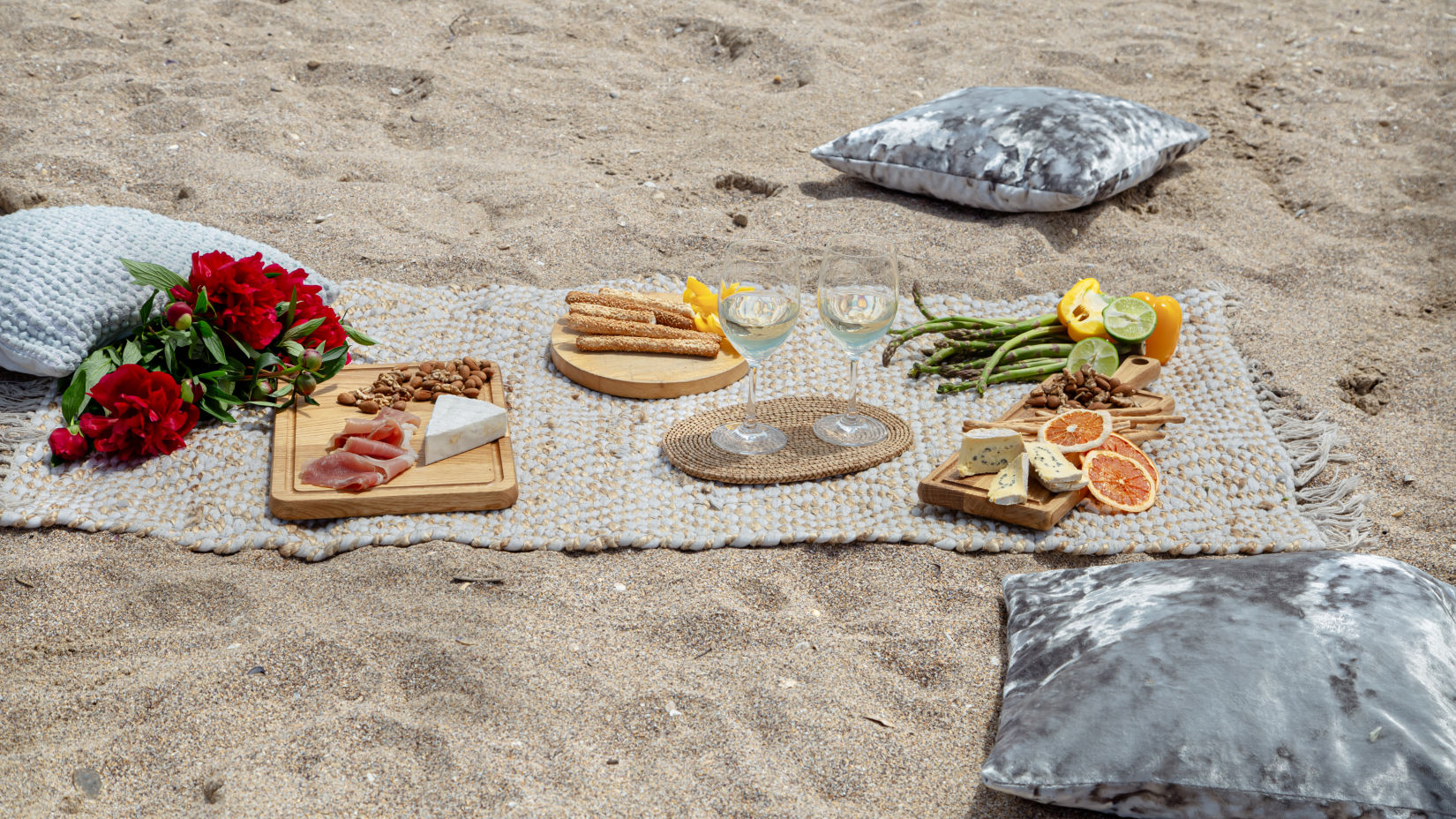 a picnic setup with food fruits and flowers placed atop a sheet which is placed on sand