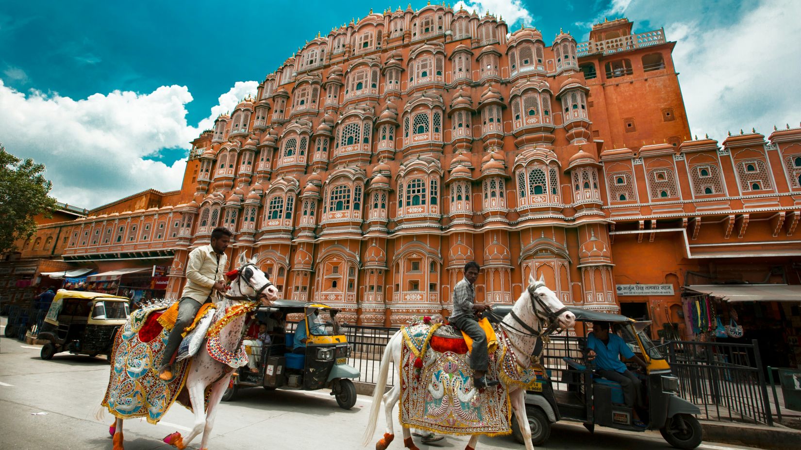 A vibrant street scene in Jaipur with ornately decorated horses and riders in the foreground and the iconic, terracotta-pink façade of the Hawa Mahal palace under a blue sky in the background.