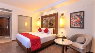A room with a clean bed, sofa, chair, table, wall painting, and lamps - The Saibaba Hotel