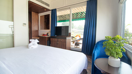 A hotel room with a large bed, blue upholstered chairs, wooden furniture, and a balcony overlooking an outdoor area.