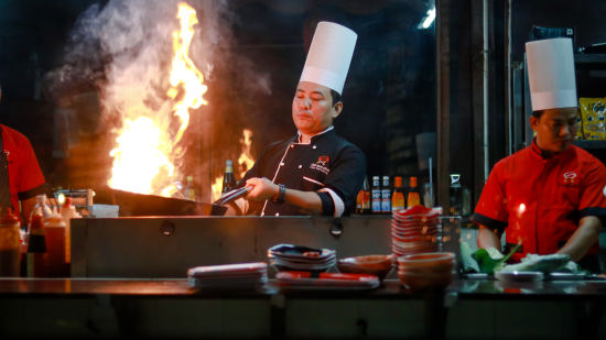 an Asian chef flambeing a dish at Buddha Garden with two other chefs next to him standing behind a pile of plates - Symphony Palms Beach Resort And Spa