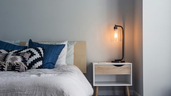 A cozy bedroom corner with a stylish bedside lamp, a modern nightstand, and a bed adorned with patterned and blue cushions.