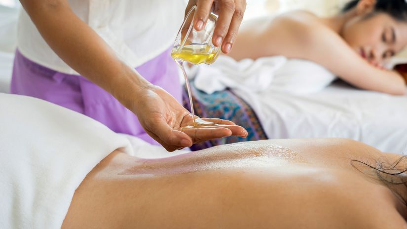 Image of a masseuse pouring oil on her palm while a woman is lying down in front of her