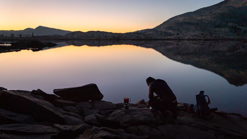 sunrise view in front of a wide lake surrounded by a mountain and a man resting after a trek