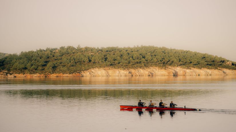 A group of rowers in a long, thin boat glide across a quiet lake against the backdrop of a hill covered in trees with ease -Fort JadhavGADH