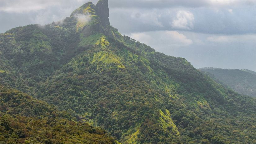  A lush green mountain peak shrouded in mist under a sky partly cloudy with sunlight piercing through- Fort Jadhav GADH