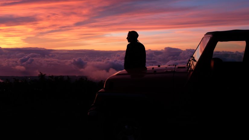 a person sitting on the hood of a car looking at the different hues in the sky