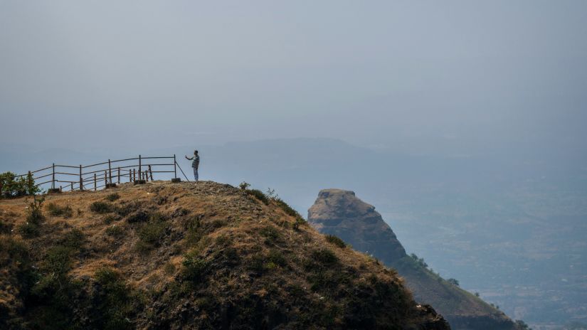 A person stands at the edge of a scenic overlook on a mountain with a view of distant peaks