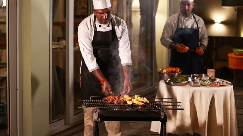 a chef grilling food on a barbecue while another chef can be seen prepping the food in the background - Lamrin Norwood Green, Palampur