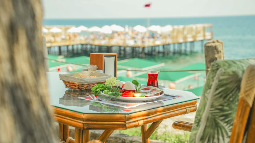 A table with food and drink items on it and comfortable chairs in view with a pier in the background