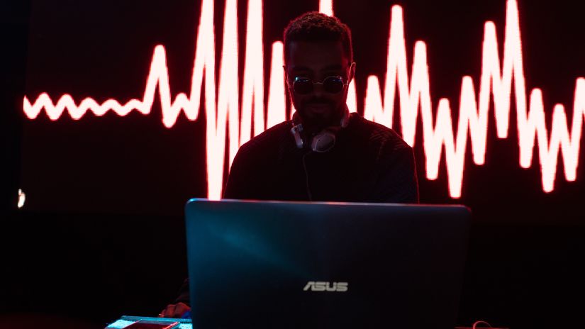 a DJ wearing a sunglass and a headphone playing his tracks and standing on the stage with his laptop