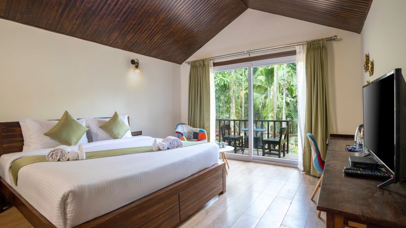 A hotel bedroom with a large bed with a green runner, a television and a balcony with the view of the garden - Stone Wood Nature Resort, Gokarna