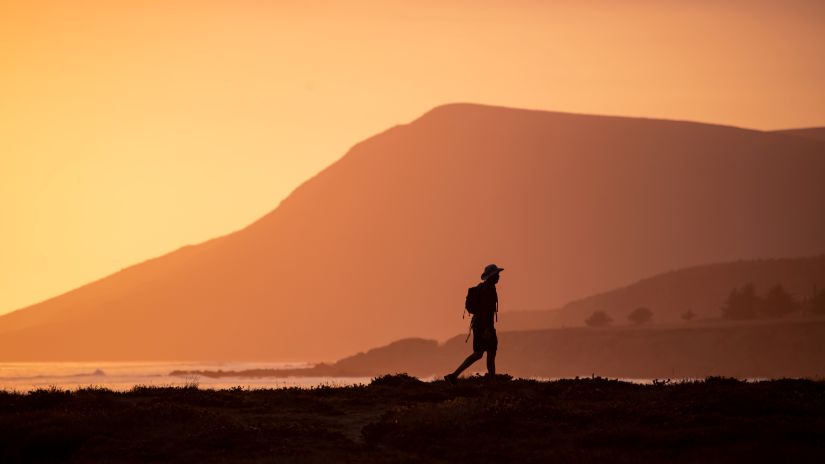 a man captured with a hiking backpack can be seen walking besides the shore during sunset with mountains as a backdrop