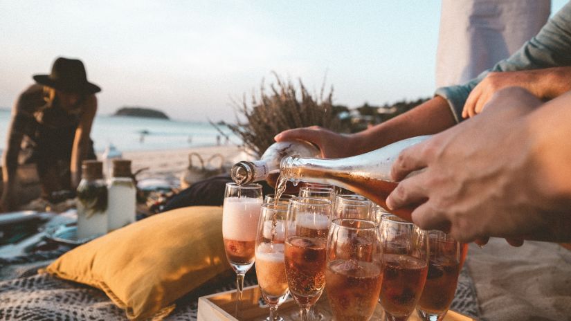 Image of a hand holding a bottle of aerated beverage that is being poured into numerous glasses placed on a rug