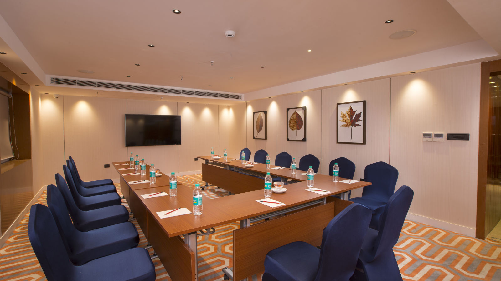 A formal boardroom with blue chairs around a long table, a flat-screen TV on the wall and framed leaf prints - Hotel Southend by TGI - Bommasandra, Bangalore