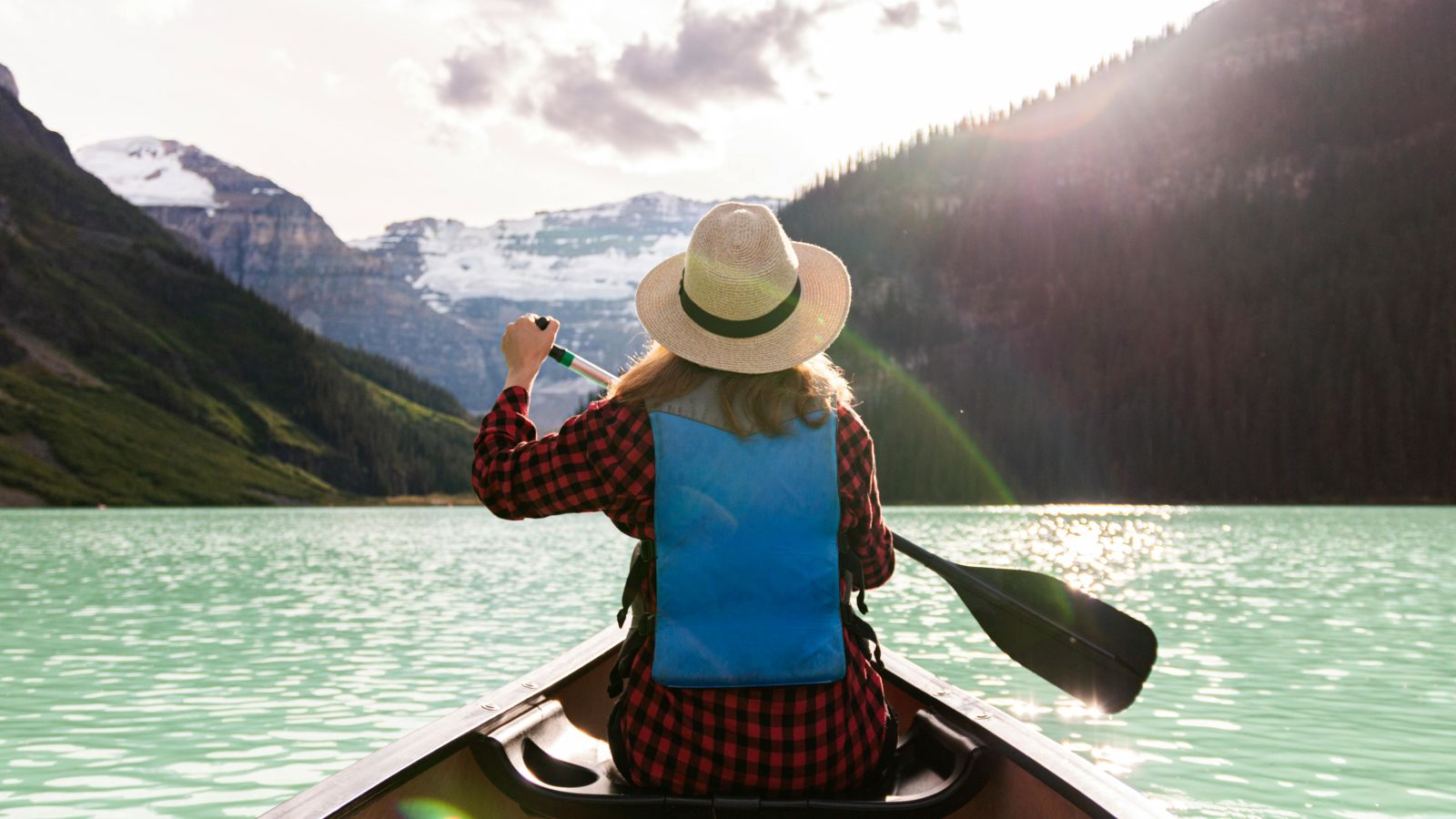 A person in a hat and life jacket is canoeing on a glacial lake, with majestic snow-capped mountains in the background - Kumarakom 